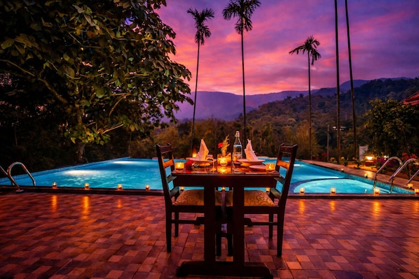 Une table à manger dressée pour deux personnes, avec vue sur une piscine et des montagnes en arrière-plan.