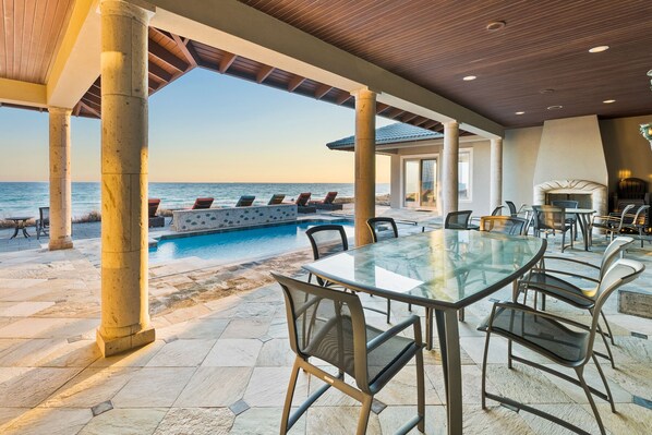 An outdoor pool area with a view of the sea, surrounded by stone pillars and furnished with lounge chairs and a glass table.