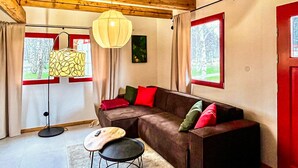 A cozy living room with a brown sofa, red and green cushions, a wooden ceiling, and large windows with curtains.