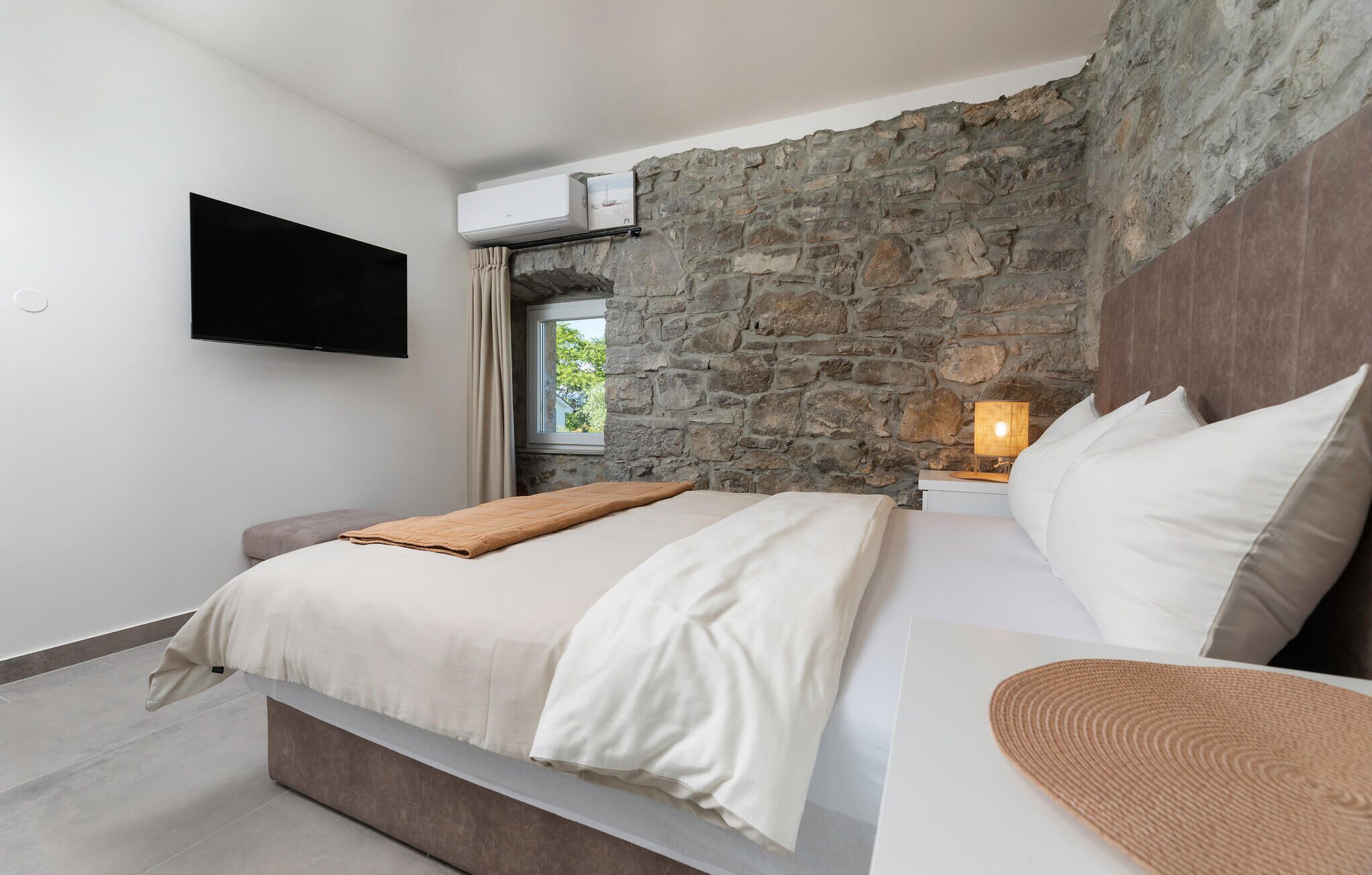 A bedroom with a stone wall, a flat-screen TV, a bed with white linens, and a bedside lamp.