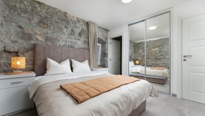 A modern bedroom with a stone wall, a large bed with white bedding, a bedside table with a lamp, and a sliding glass door leading to another room. - Gorgeous home in Lakmartin (Lakmartin)