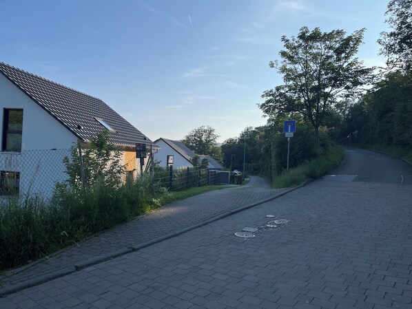 A paved road with a blue traffic sign, a white building with a tiled roof, and a tree-lined path.