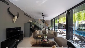 A modern living room with a large flat-screen TV, a sofa, a coffee table, and a glass door leading to a pool area.