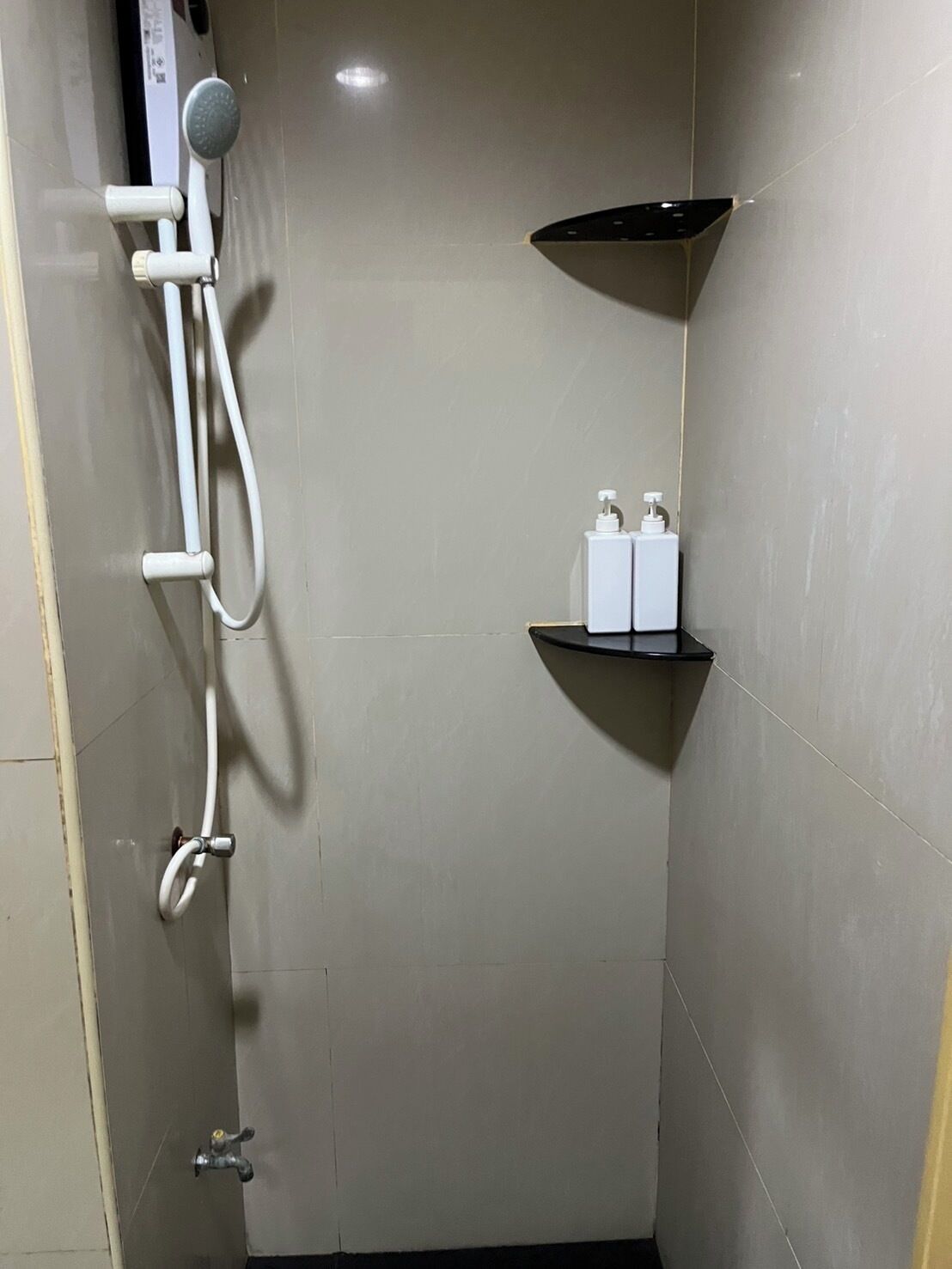 Une petite salle de bain carrelée, équipée d’une douche, d’un pommeau de douche amovible, d’un flexible de douche, d’un distributeur de savon et d’une petite étagère.