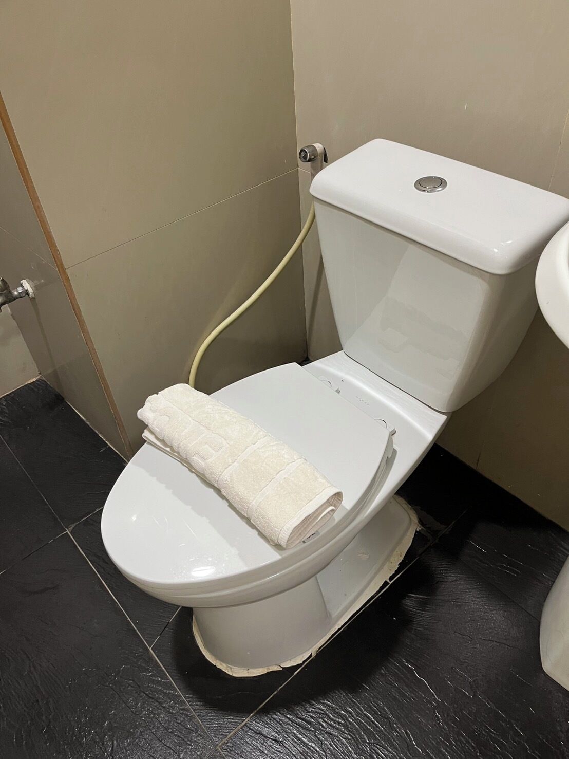 En vit toalett med en handduk på sitsen, en beige vägg och ett svart kaklat golv.