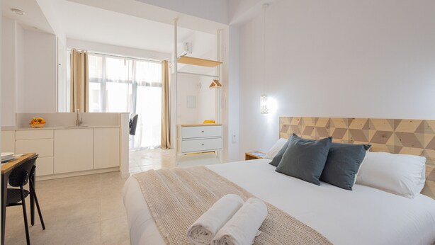 A modern hotel room with a large bed, a desk, and a chair. There is a window with curtains, a wooden headboard, and a bedside table with a lamp. - The Sorolla Apartment V | AC & WiFi | 4 Guests (Valencia)