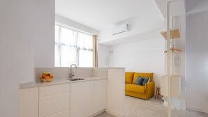 A modern kitchen with white cabinetry, a sink, and a window with curtains. There is a yellow sofa in the background. - The Sorolla Apartment V | AC & WiFi | 4 Guests (Valencia)