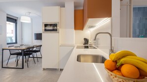 Une cuisine moderne avec un plan de travail blanc, un évier en acier inoxydable et un panier rempli d’oranges et de bananes.