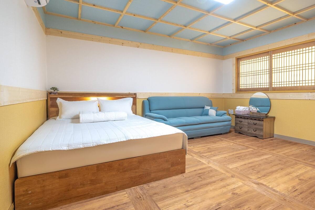 A bedroom with a wooden bed, a blue sofa, a wooden nightstand, and a window with wooden blinds.