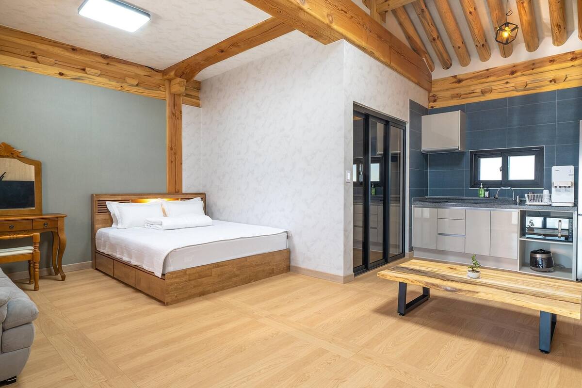 A bedroom with a bed, a wooden desk, and a bench. There is a kitchen with a refrigerator, a microwave, and a sink.