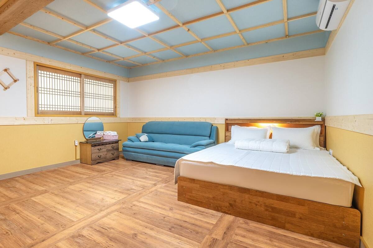 A bedroom with a wooden ceiling, a bed, a blue sofa, a small table with a mirror, and a window with bars.