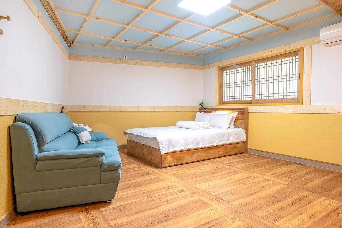 A room with a wooden floor, a bed with a white mattress and pillows, a blue sofa, and a window with wooden blinds.