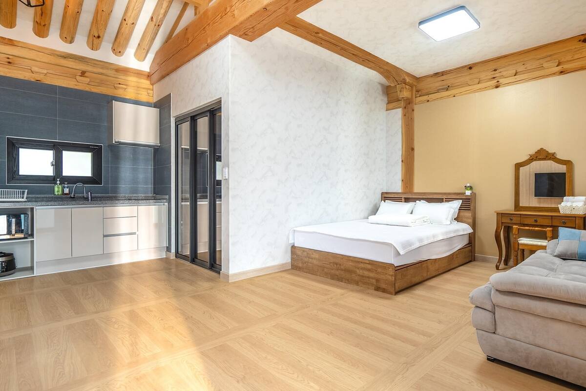 A modern hotel room with a wooden ceiling, a bed, a sofa, a small table, and a kitchen area with a sink and cabinets.