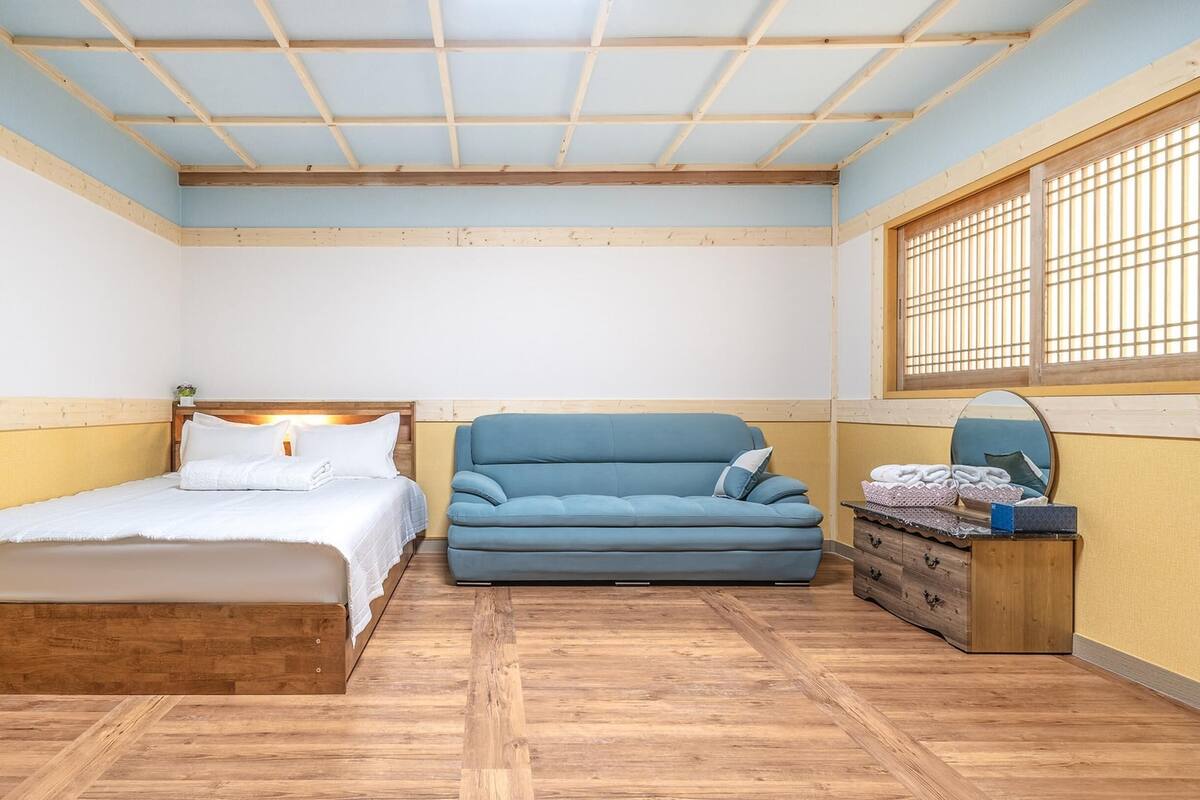 A bedroom with a bed, a blue sofa, a wooden dresser, and a window with blinds.