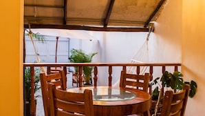 Een ronde houten tafel met stoelen in een overdekte buitenruimte met uitzicht op een tuin.