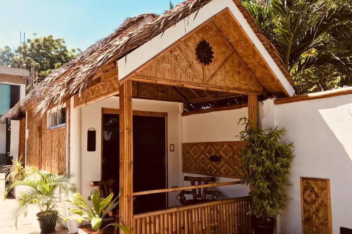 A thatched-roof structure with a wooden porch, potted plants, and a bamboo fence.
