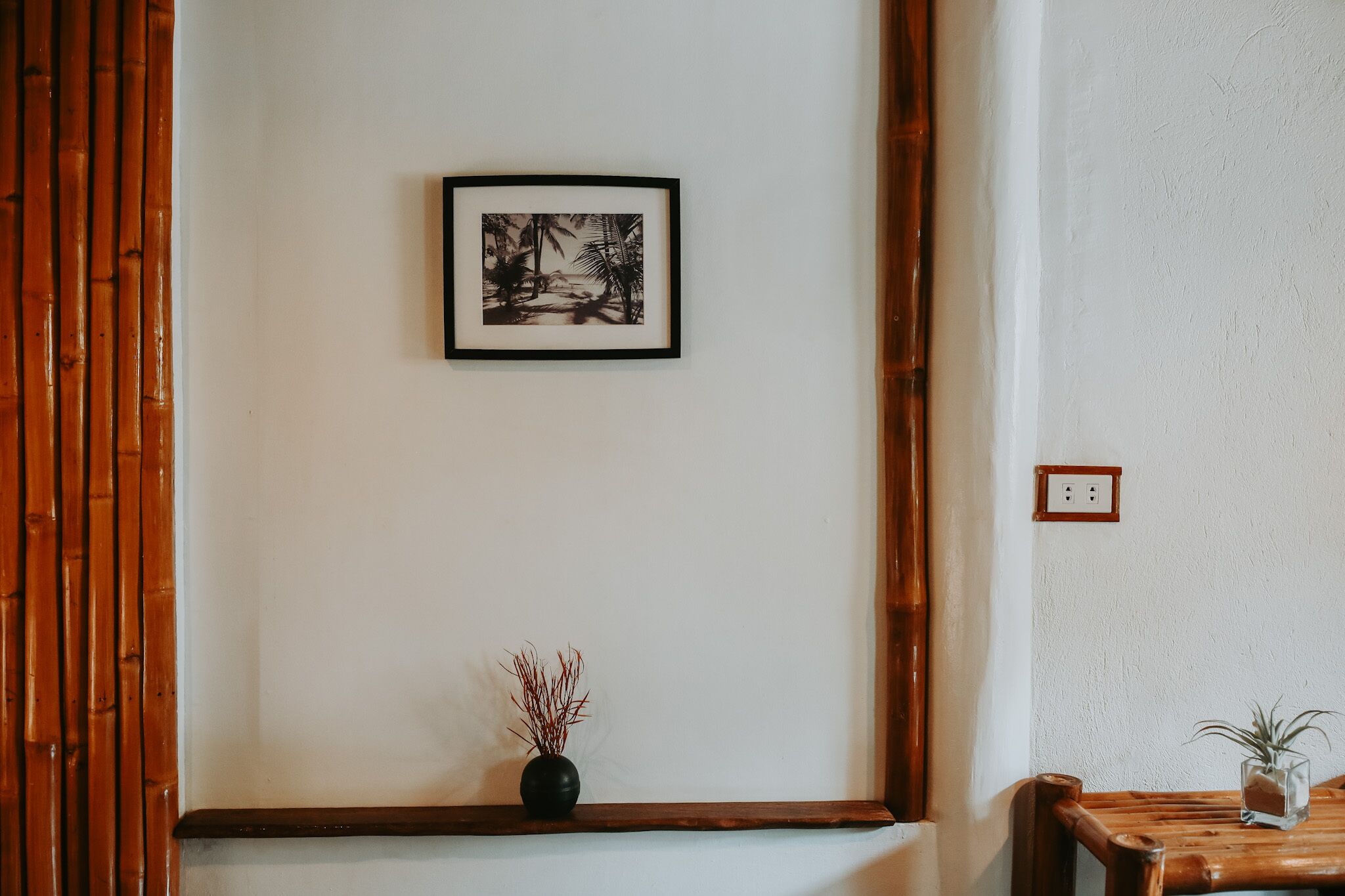 Habitació amb una banca de fusta, una taula petita amb una planta, un quadre amb marc a la paret i una pantalla de bambú.