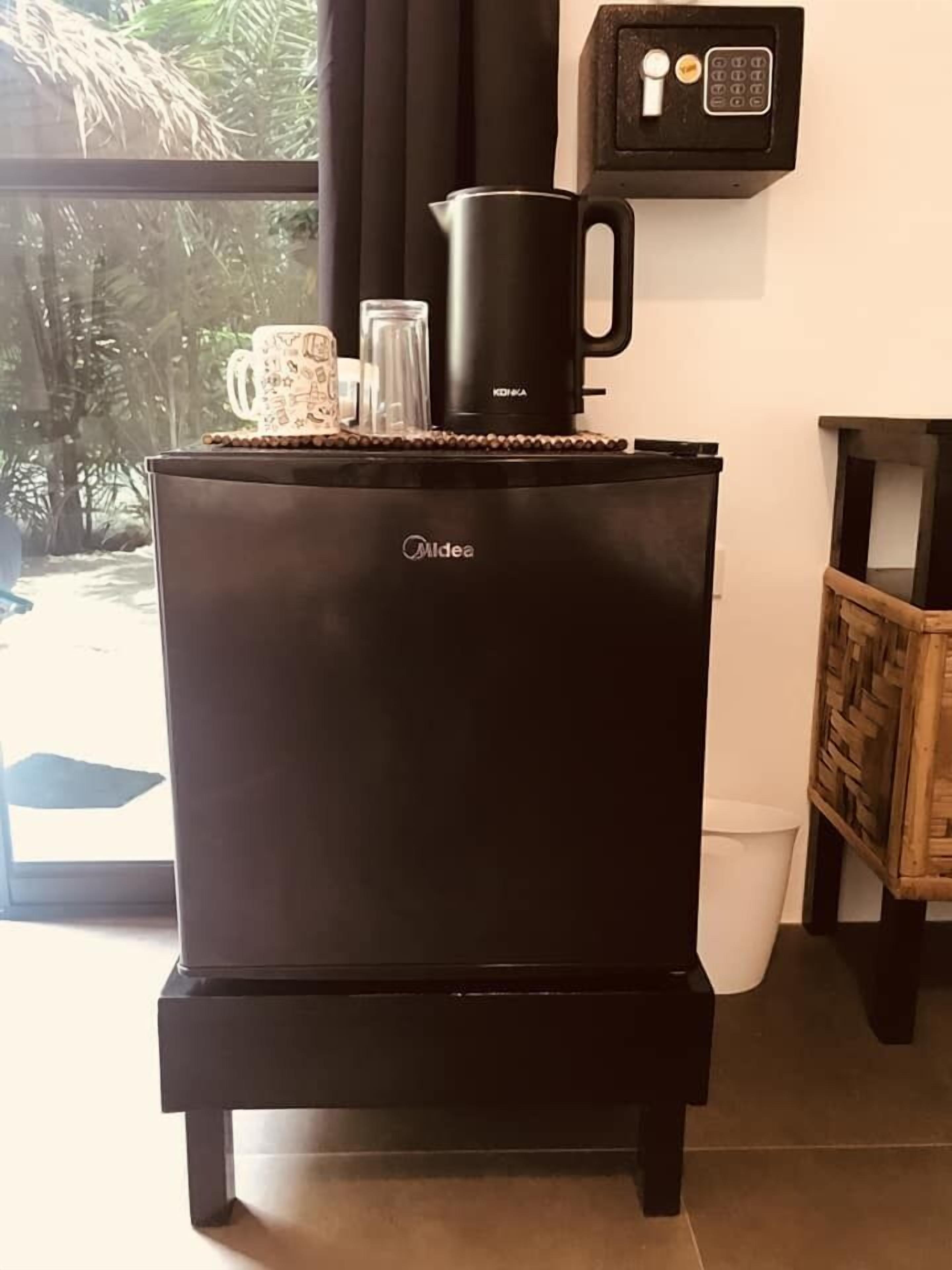 A black Midea refrigerator with a coffee maker on top, a small shelf with a glass and a mug, and a window with a view of greenery.