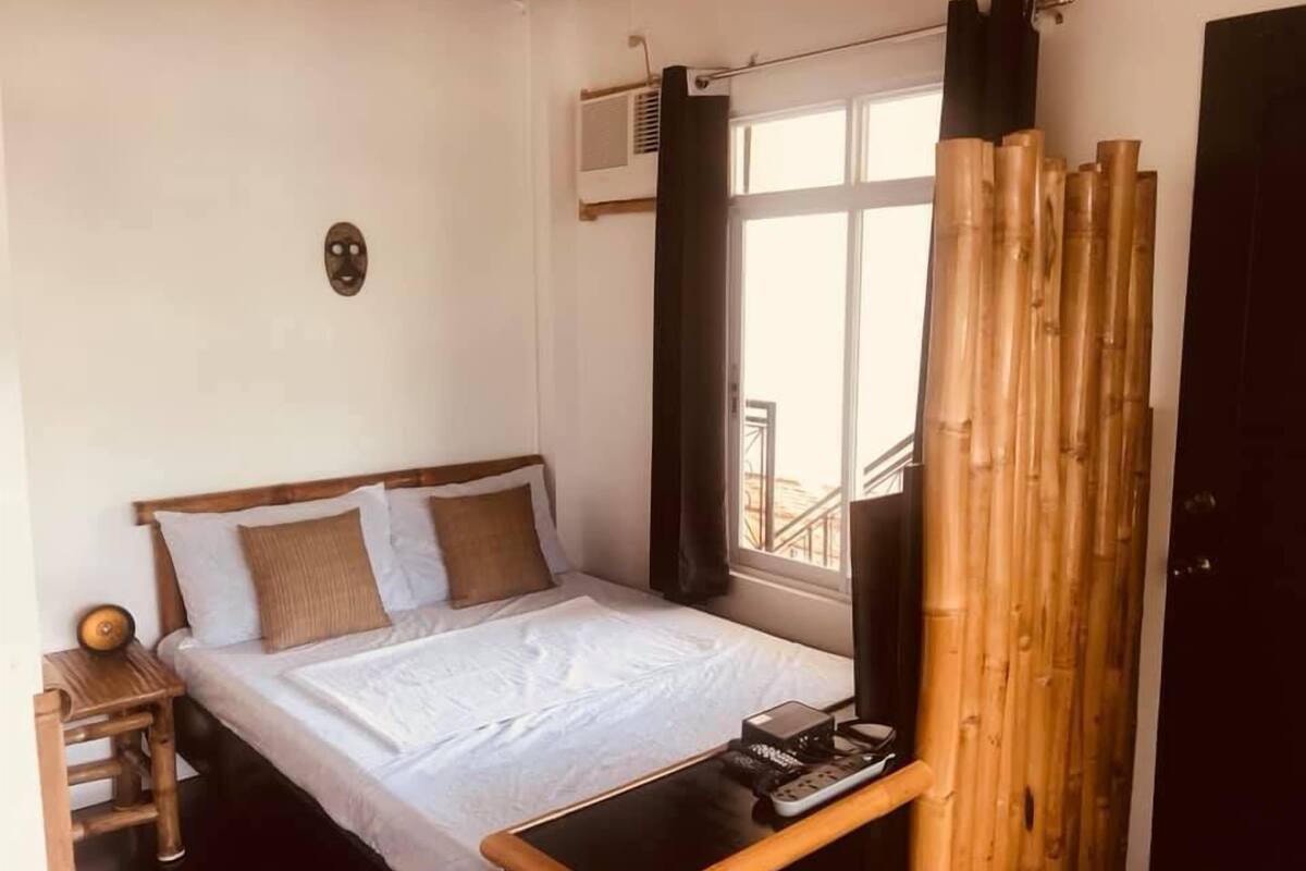 A bedroom with a bed, a bedside table with a remote control, a wooden chair, and a window with curtains.