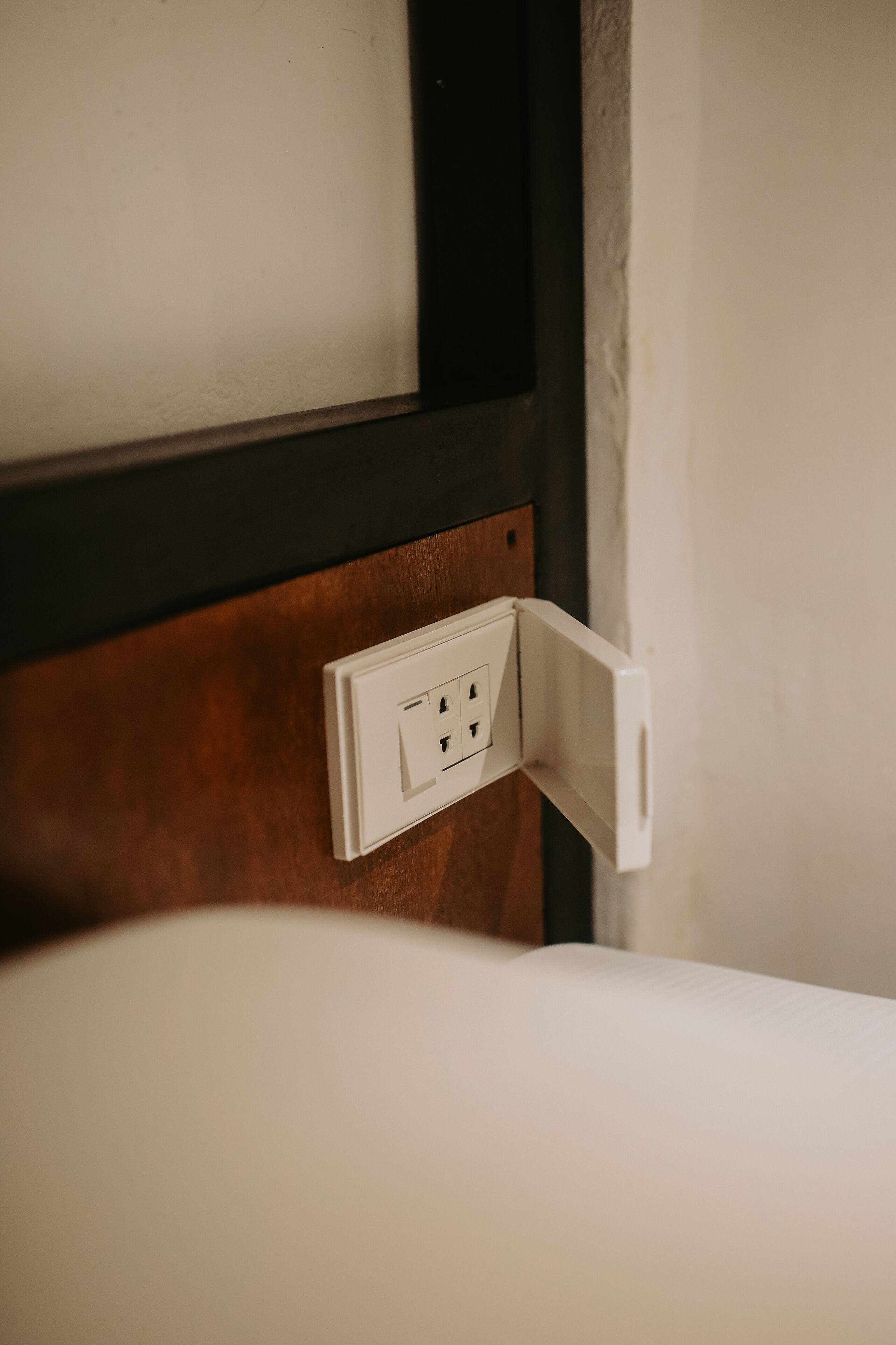 A white electrical outlet on a wooden panel, likely in a hotel room.