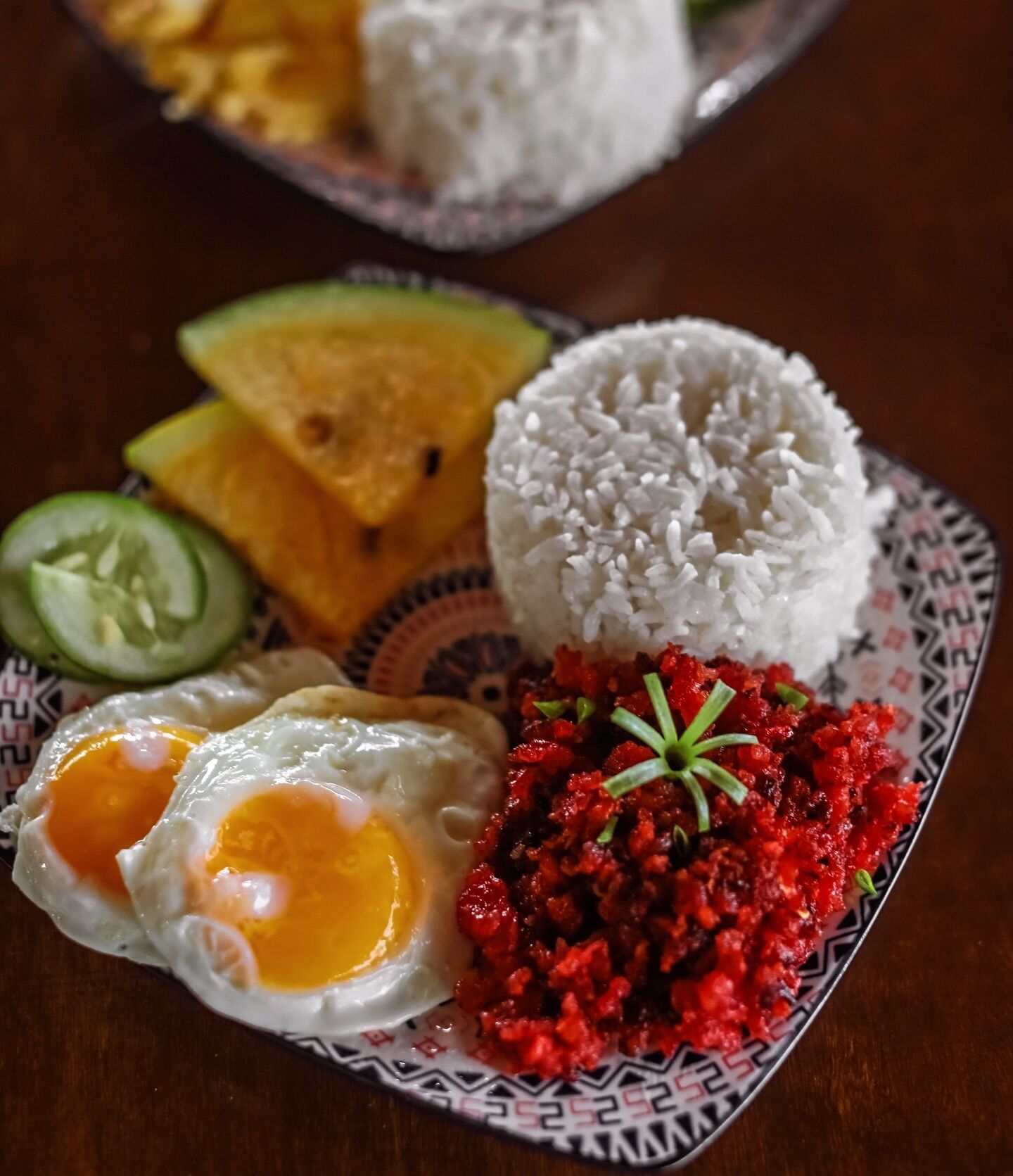A plate with white rice, two fried eggs, a red vegetable dish, slices of cucumber, and a slice of yellow fruit.