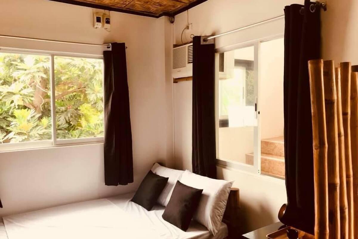 A single bed with white bedding and two pillows, a window with a view of greenery, and a bamboo curtain.