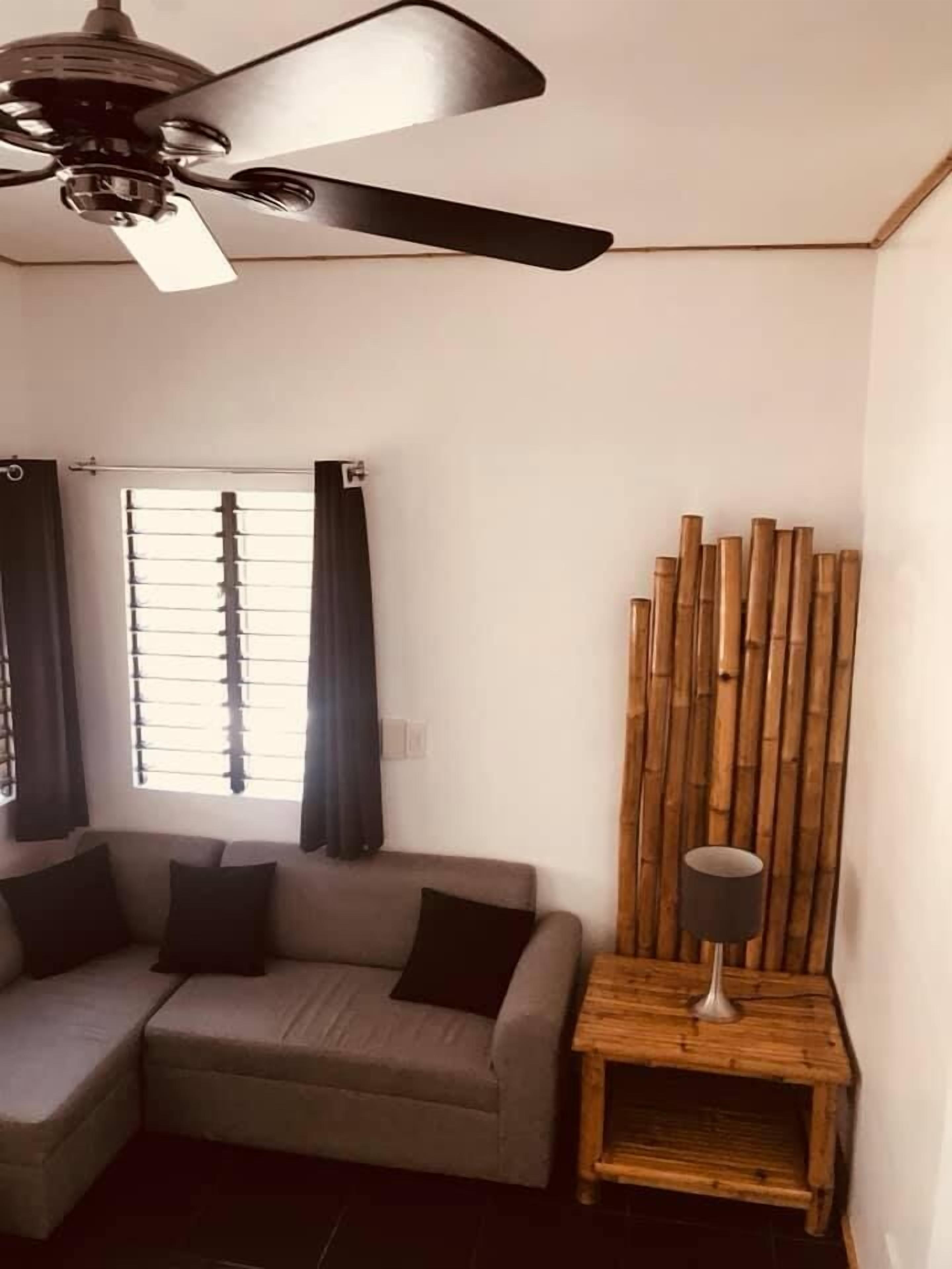 Una sala de estar con ventilador de techo, un sofá, una ventana con persianas y un adorno de pared de madera.