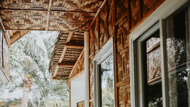 A wooden exterior with a woven ceiling, large windows, and a view of palm trees.