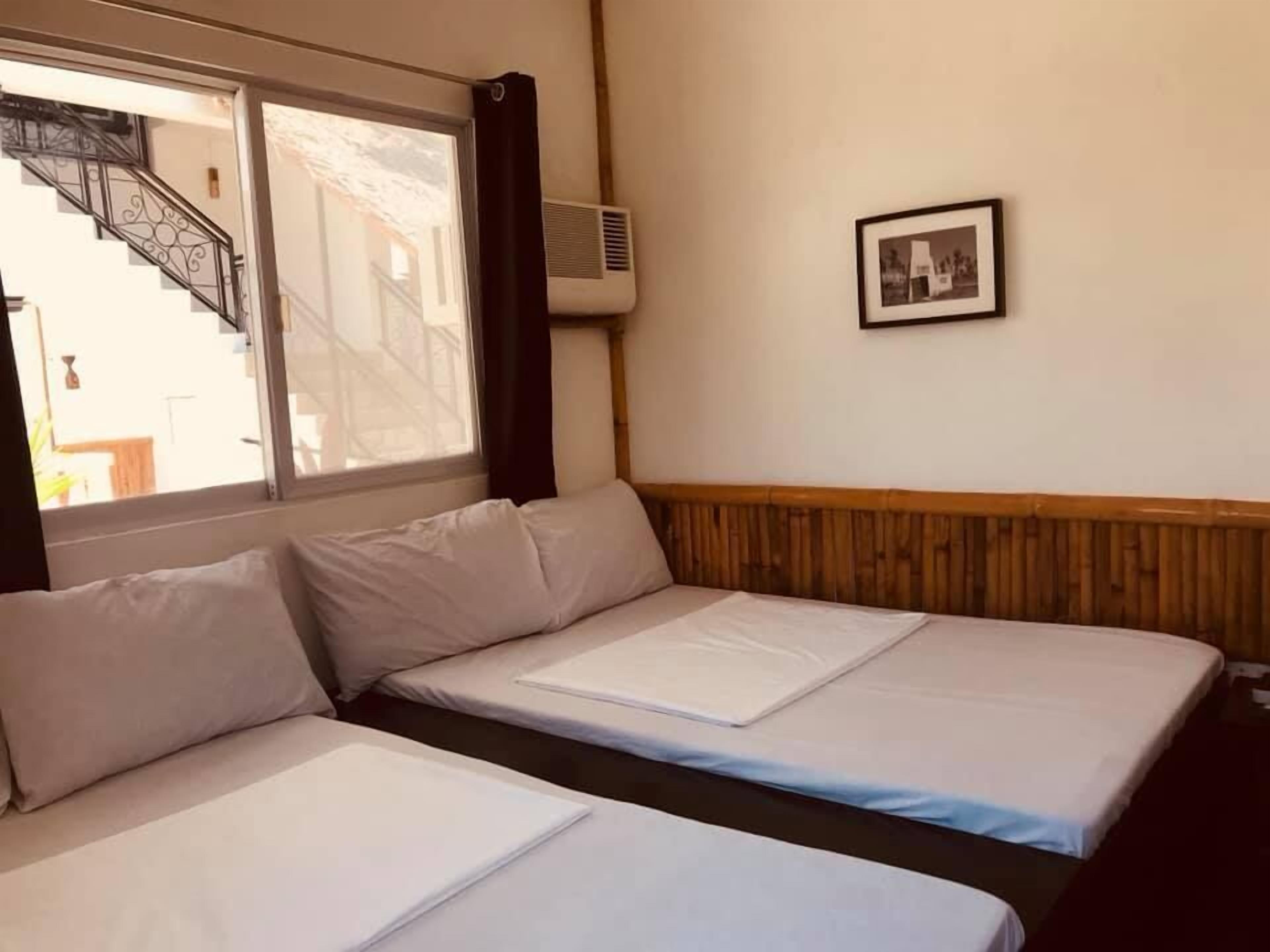 Une chambre avec deux lits, une fenêtre donnant sur un escalier, un appareil de climatisation et un tableau encadré au mur.