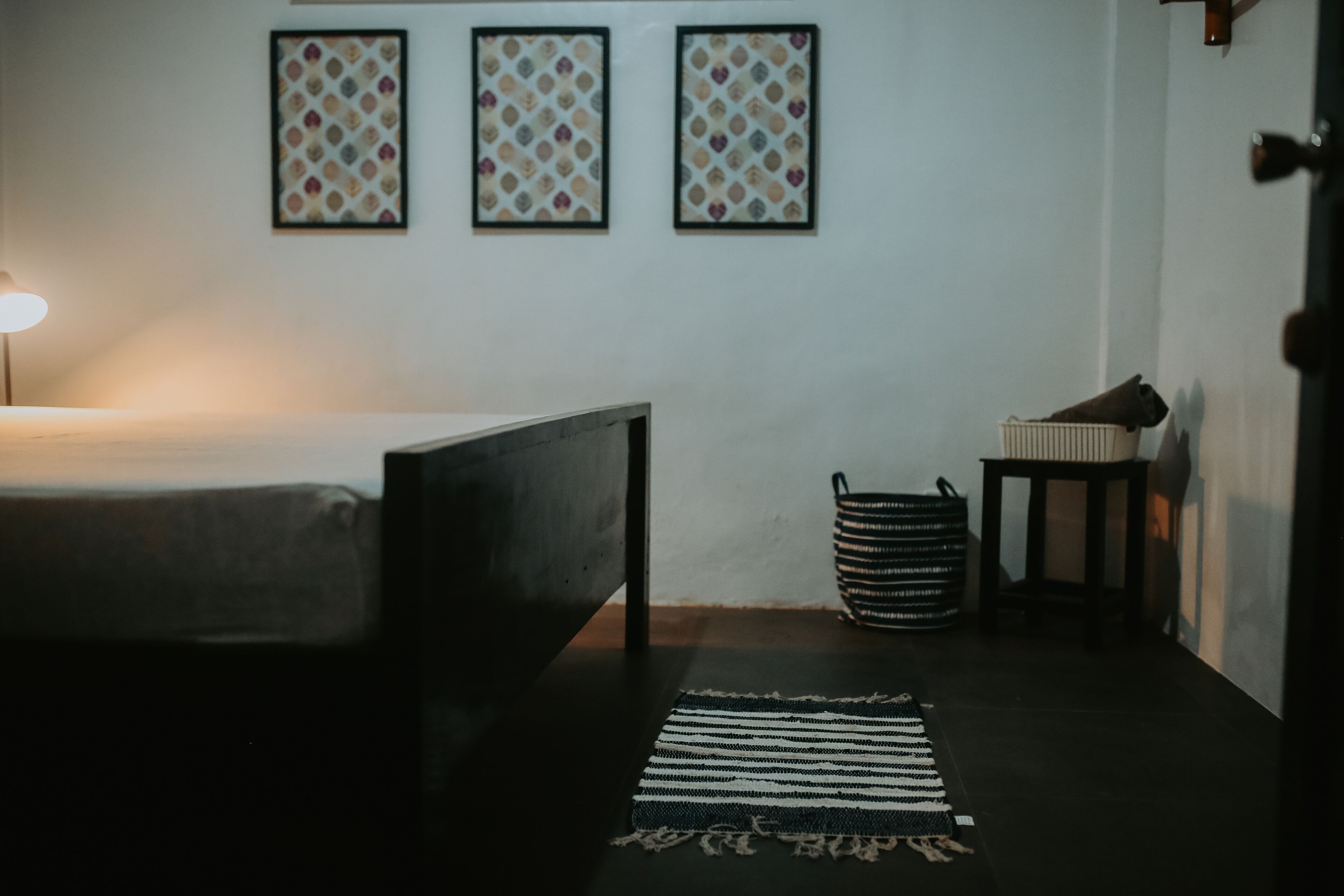 A bedroom with a bed, a bedside lamp, a small table, and a trash bin. There are three framed pictures on the wall.