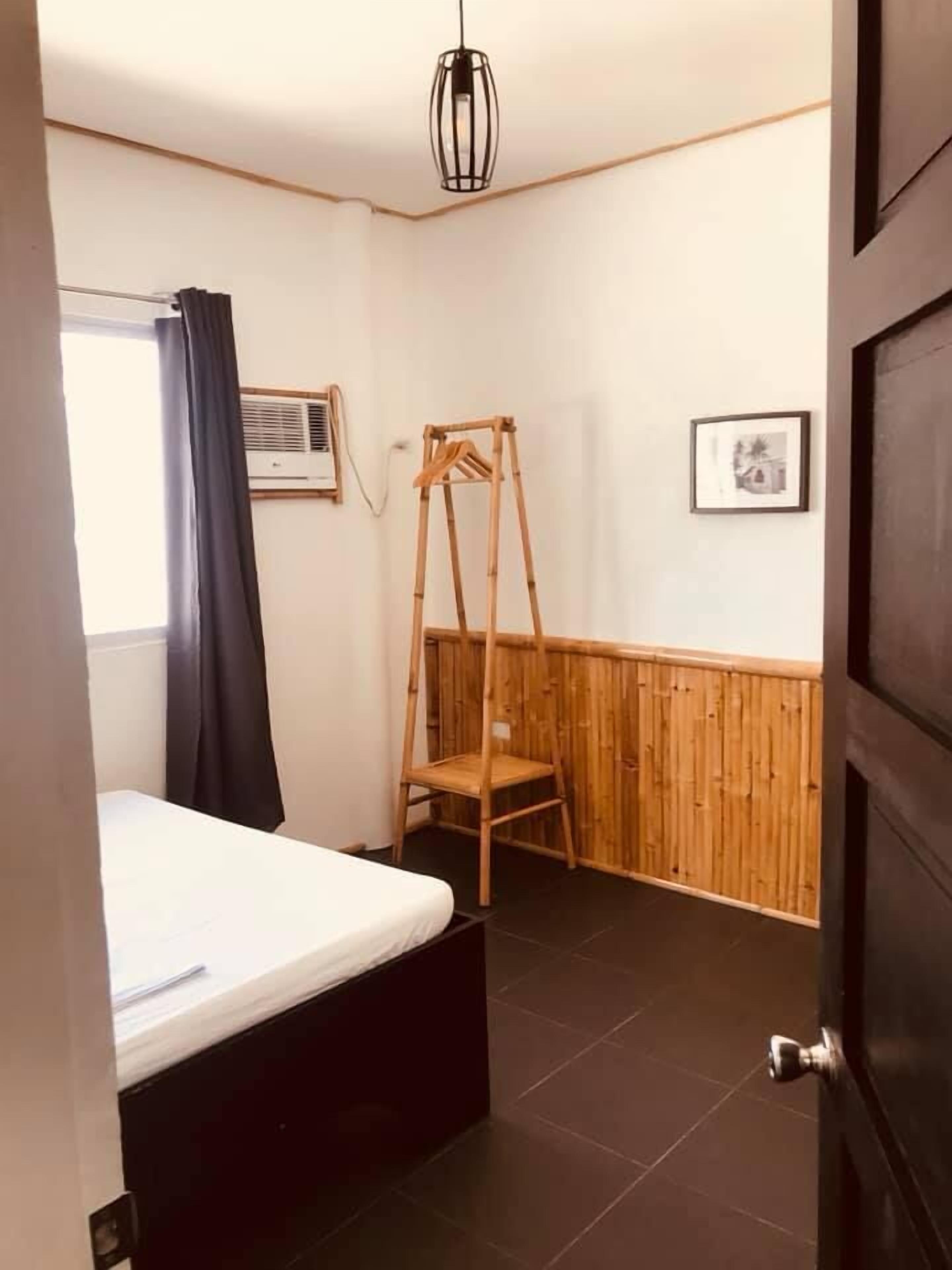 A small, well-lit room with a bed, a wooden chair, a wall-mounted air conditioner, and a door.