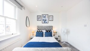 Ein modernes Schlafzimmer mit einem großen Bett, zwei Nachttischen mit Lampen, einem Standventilator und einem Kunstwerk an der Wand.