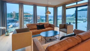 A modern living room with a sofa, armchairs, and a coffee table. Large windows offer a view of a residential area and hills. - 3 bedroom lovely home in Lindesnes (Lindesnes)
