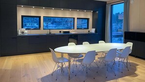 A modern kitchen with a dining area, featuring a large island, built-in appliances, and a view of a residential area through the windows. - 3 bedroom lovely home in Lindesnes (Lindesnes)