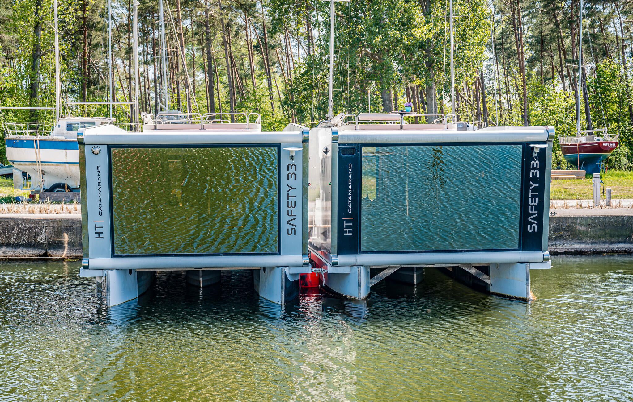 Two safety barriers labeled "Safety 33" by HT Catamarans in a water body with boats and trees in the background.