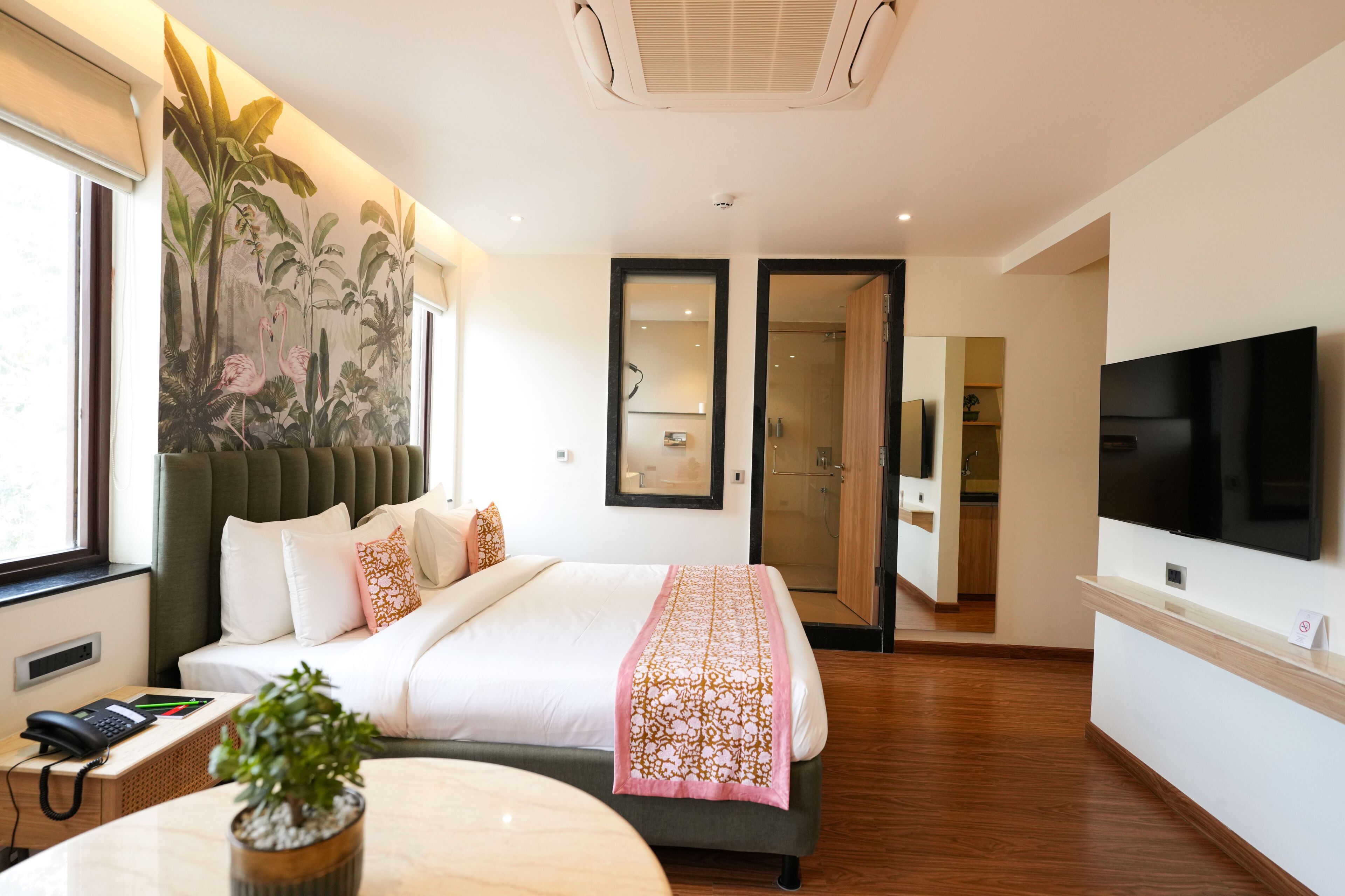 A hotel room with a large bed, a TV, a desk with a phone, a plant, and a mural on the wall.