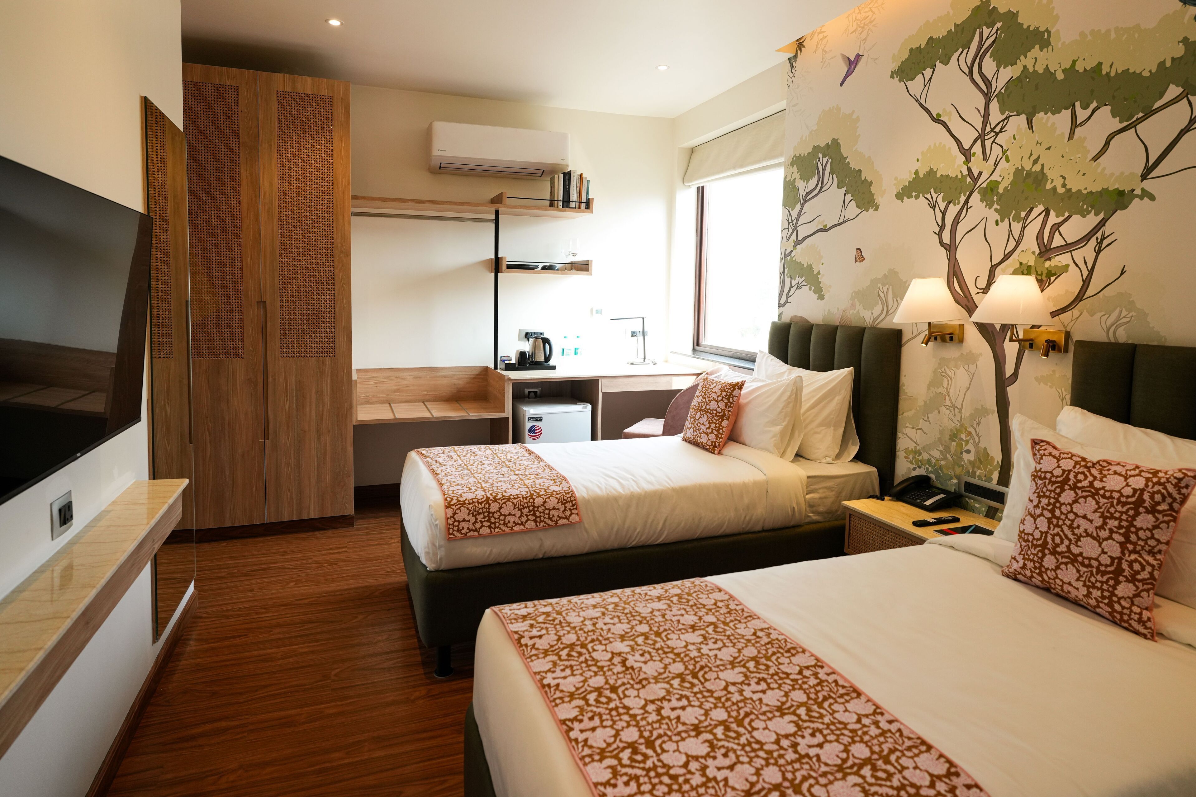 A hotel room with two beds, a desk, a TV, and a mural of trees.