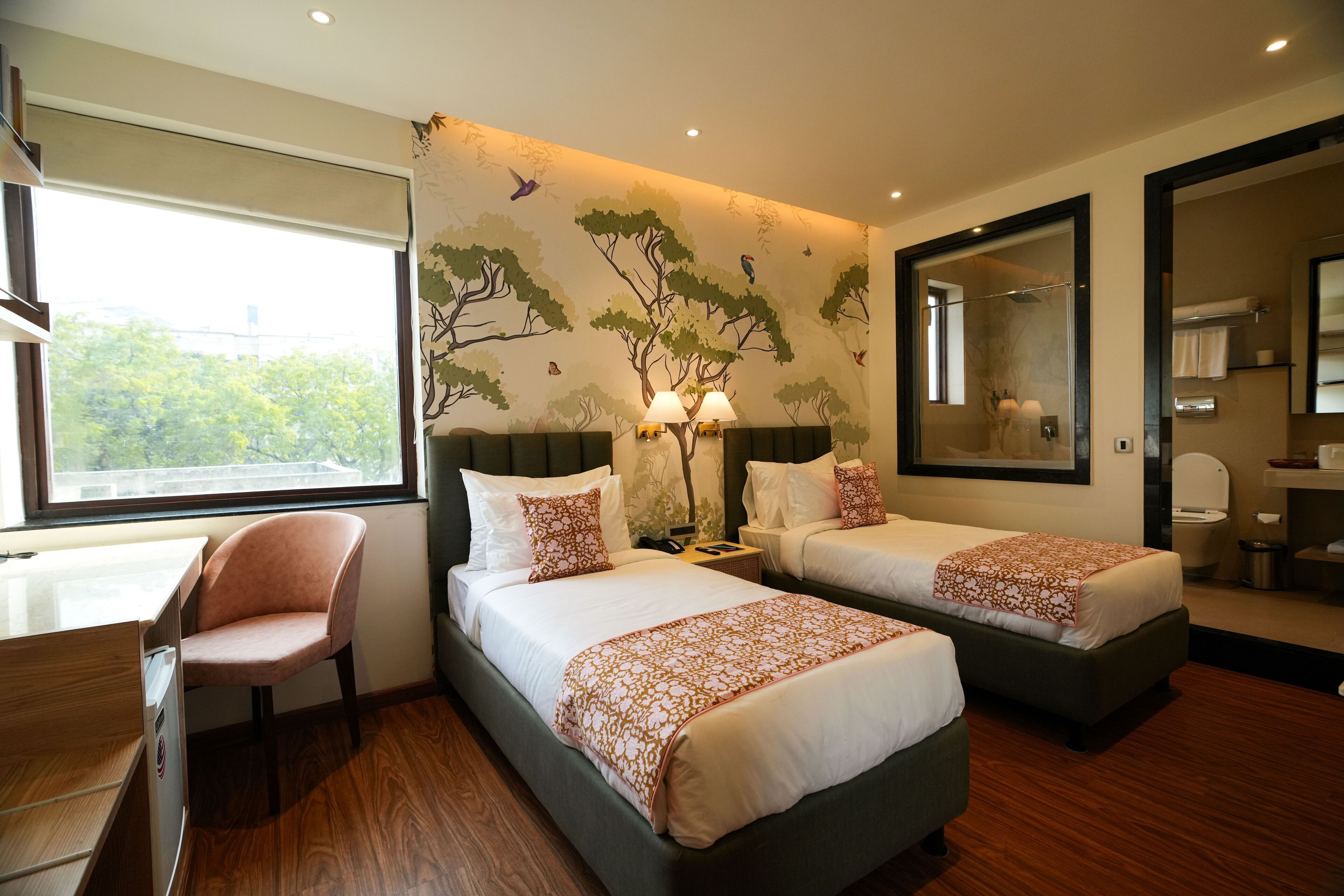 A hotel room with two beds, a desk, and a chair. There is a large window, a mirror, and a wall with a tree mural.