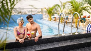 A couple by a pool with a drink, surrounded by palm trees and outdoor furniture.