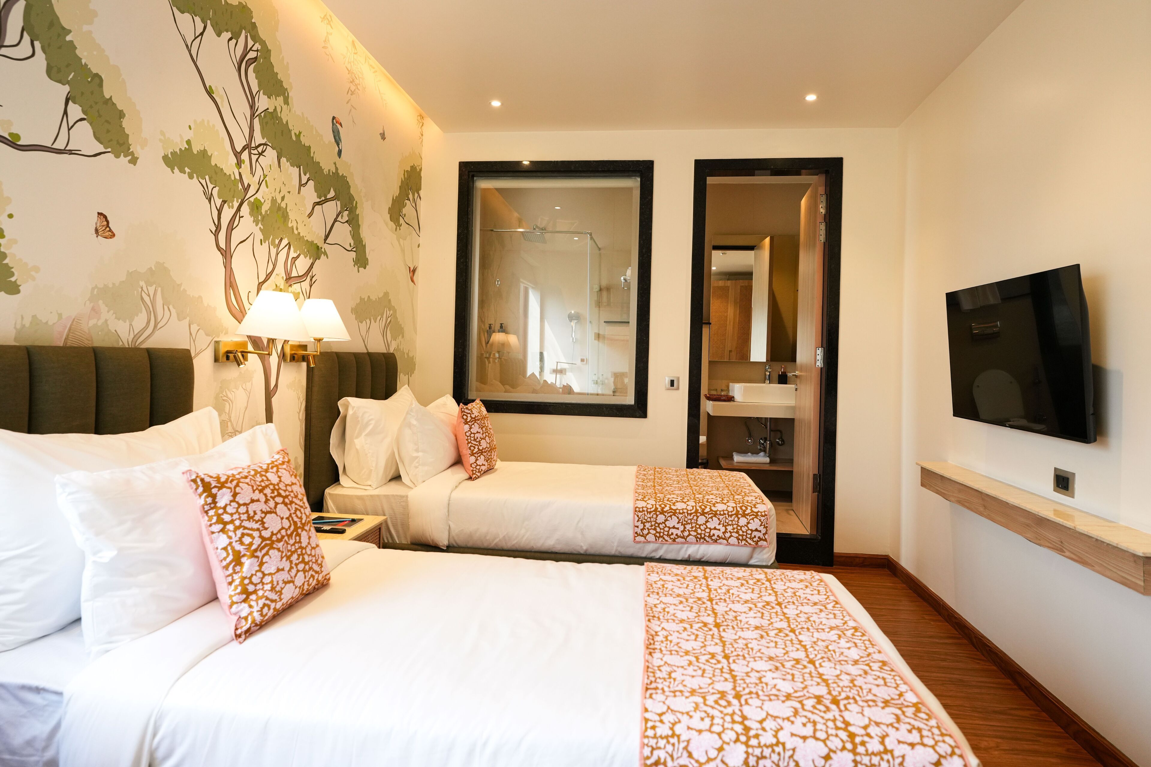 A hotel room with two beds, a TV, a mirror, and a bathroom visible through a glass door.