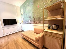 Quarto com televisão de tela plana na parede, um sofá, prateleira de vime com chaleira e papel de parede floral.