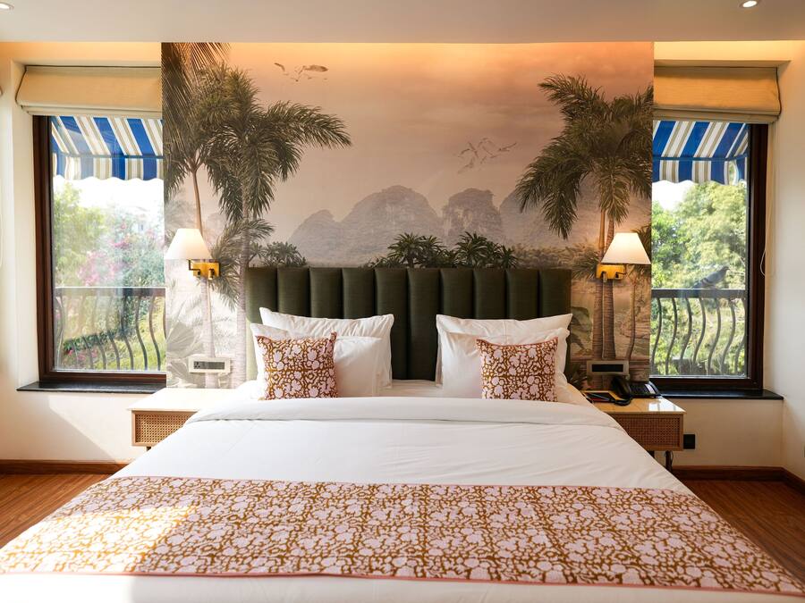 Una habitación de hotel con una cama grande, dos mesitas de noche, una lámpara de pared y vistas a palmeras y montañas a través de las ventanas.