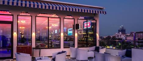 A rooftop terrace with outdoor seating and large windows overlooking a cityscape.