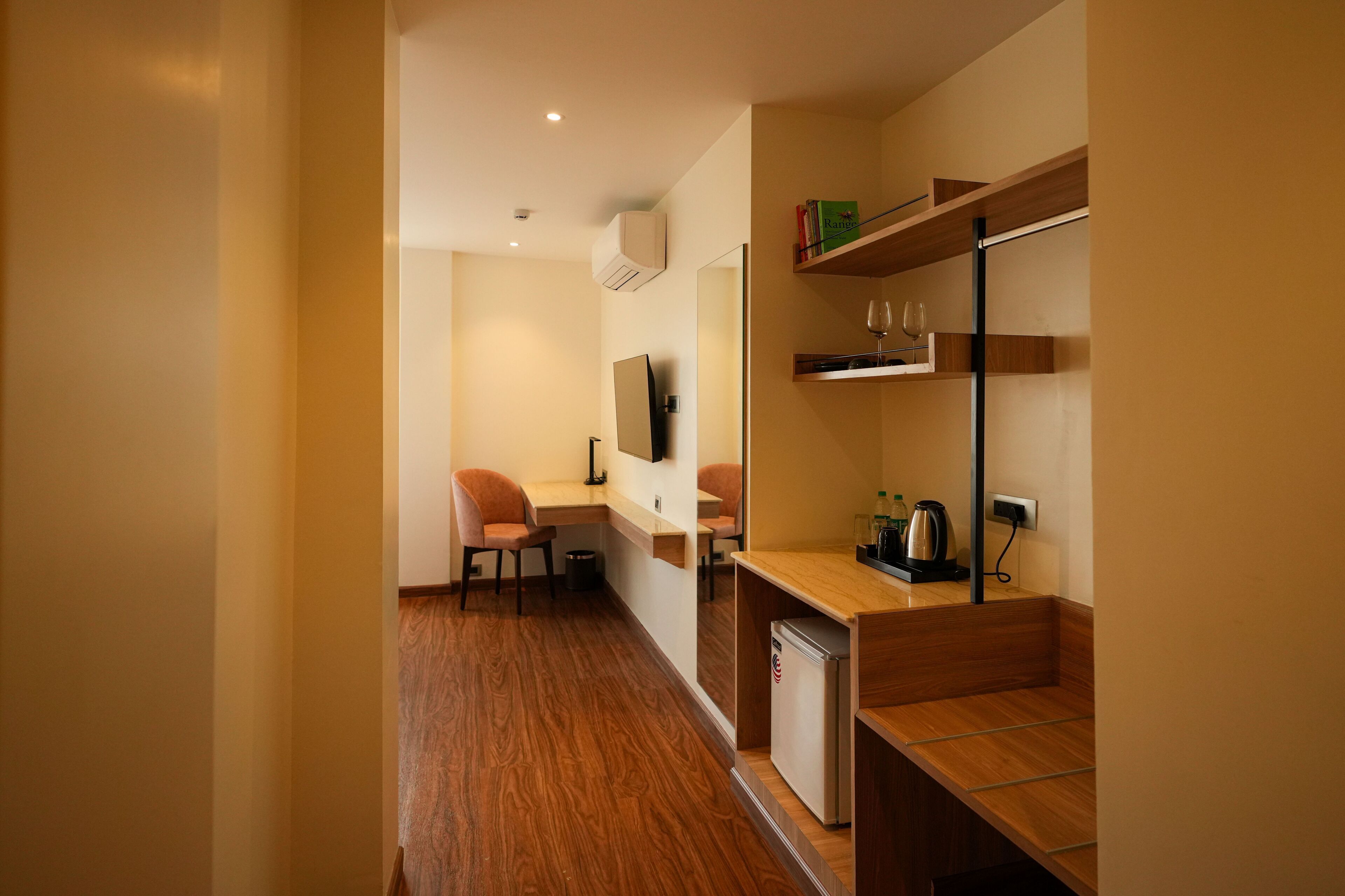 A compact hotel room with a wooden floor, a desk with a chair, a small refrigerator, a TV mounted on the wall, and a shelf with books and a wine glass.