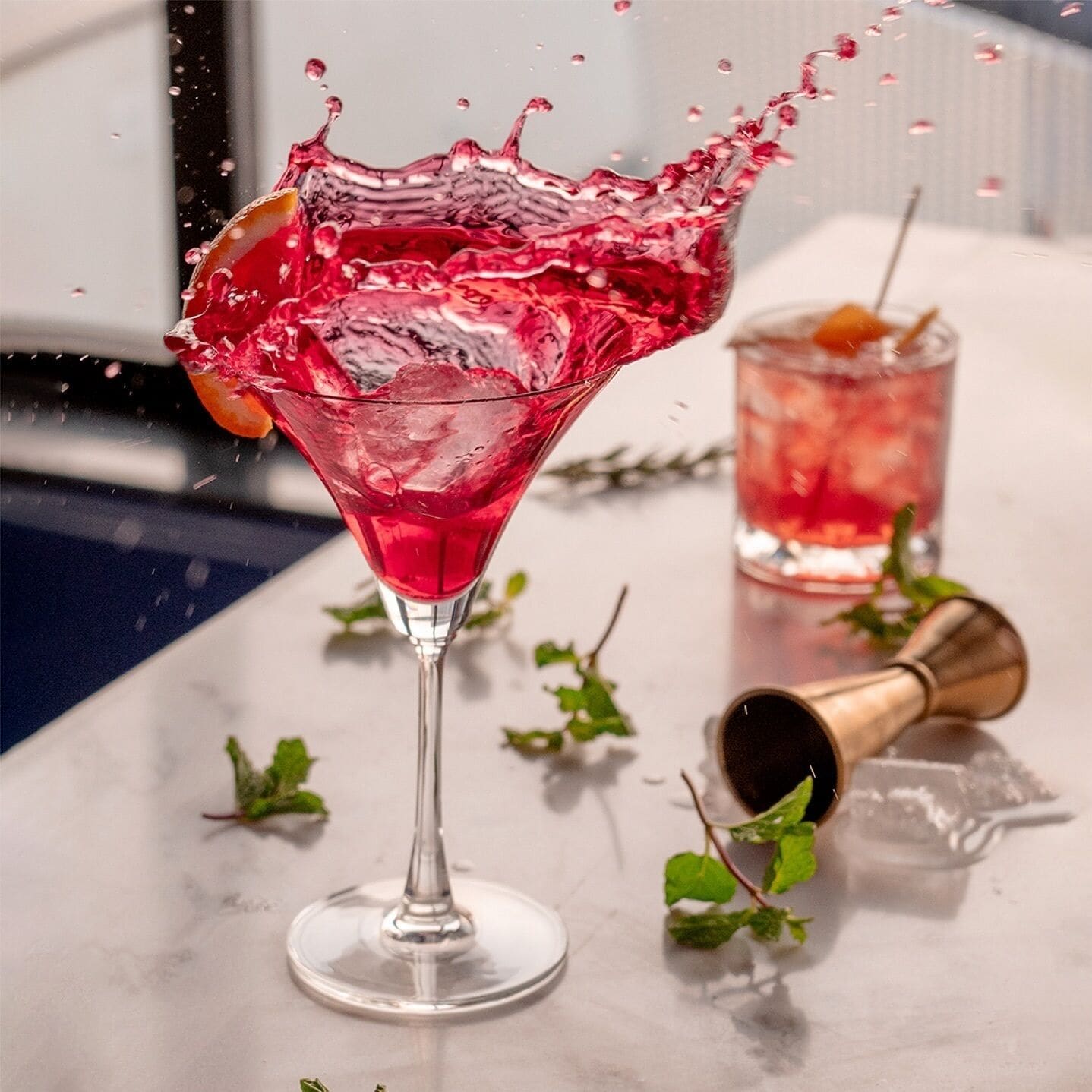 A martini glass with a splash of red liquid, a cocktail shaker, and a glass with a straw on a marble surface.