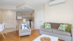 Una sala de estar con un sofá gris, un sillón blanco y una mesa de centro de madera. Se observa una cocina al fondo con armarios de madera y un refrigerador blanco.