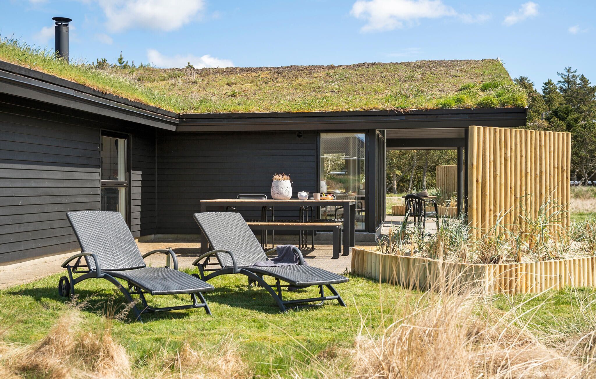 Une maison moderne avec un toit plat, un patio comprenant deux chaises longues, une table et une clôture en bois.