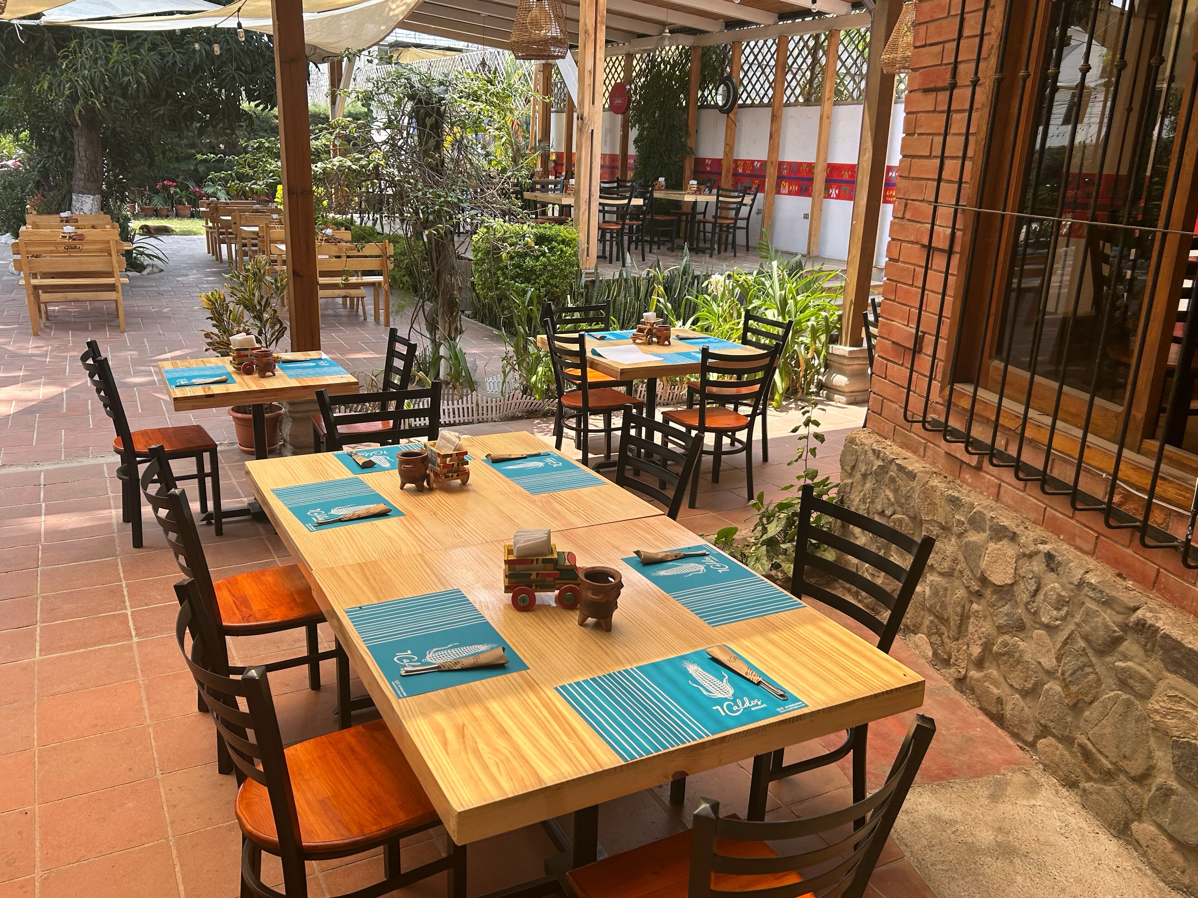 An outdoor patio with wooden tables and chairs, surrounded by potted plants and a brick wall.