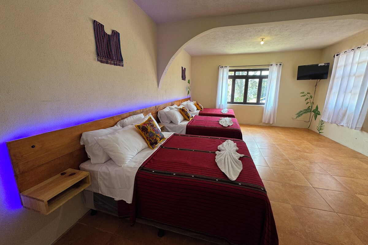 Een hotelkamer met drie bedden, een houten hoofdbord, een aan de muur bevestigde televisie en een raam met gordijnen.