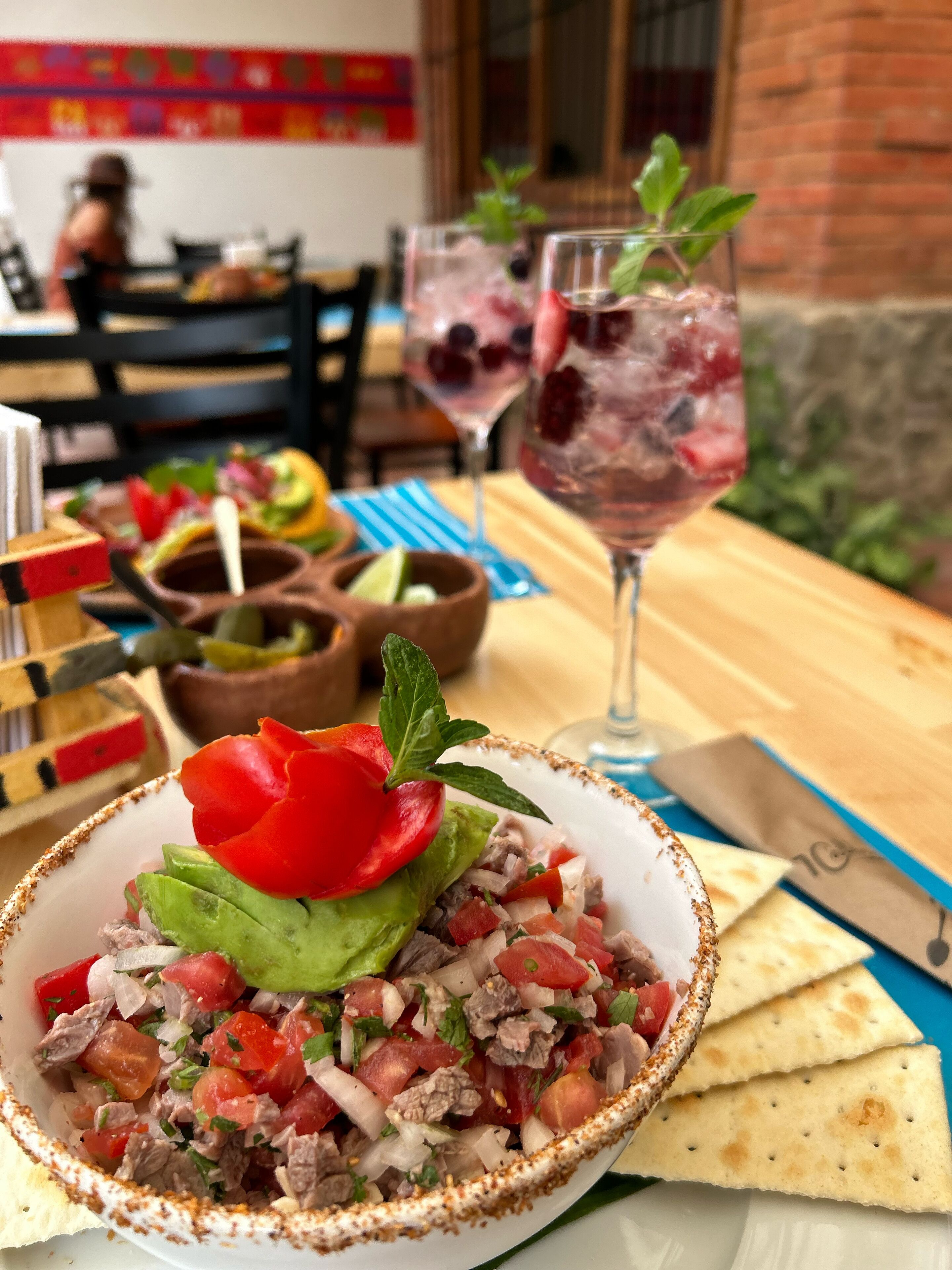 Un bol de ceviche à l’avocat et à la tomate, un verre de boisson glacée aux fruits rouges et une table en bois avec une serviette bleue et un couteau.